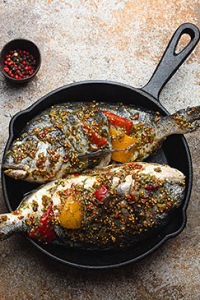 Two raw fishes dorado stuffed with vegetables and seasonings ready to be cooked laying in black cast iron frying pan top view. Healthy cooking, diet food eating concept